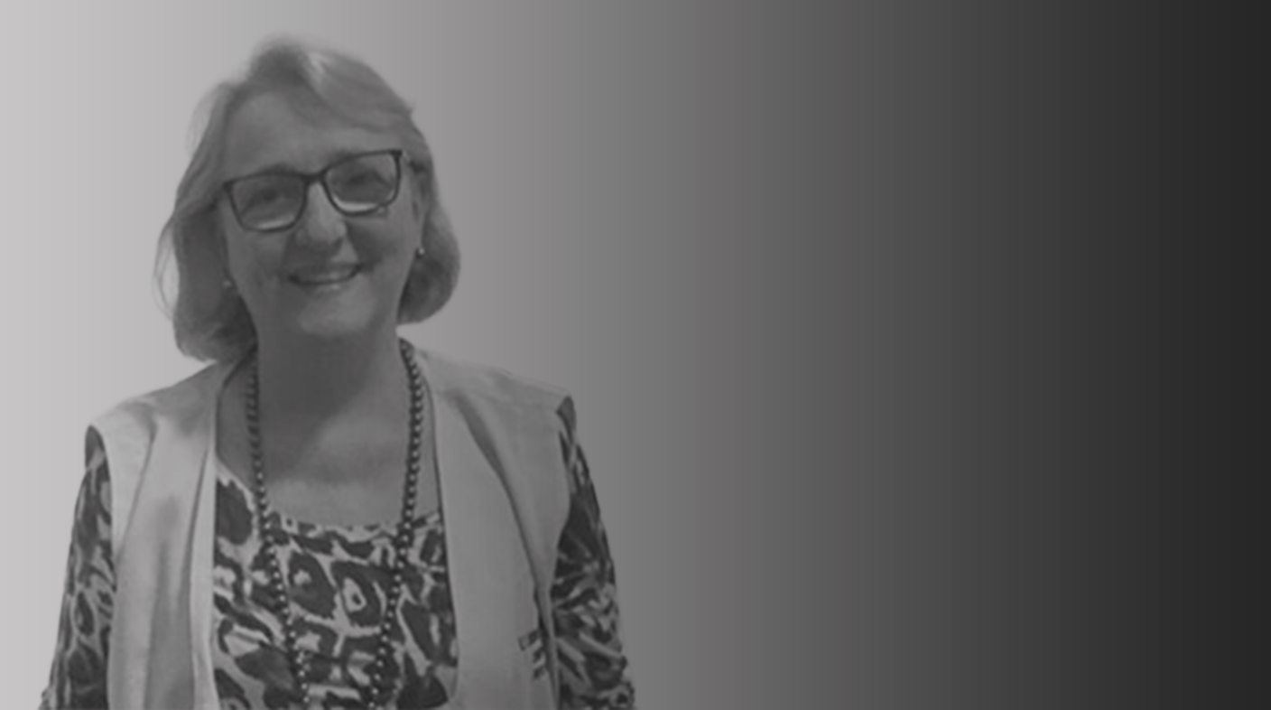 Mulher branca de fios brancos lisos e curtos, sorrindo em pé para foto. Ela veste jaleco sem mangas brancas sobre blusa estampada. Ela usa óculos e colar comprido. É a dra. Bárbara Hellebrekers.