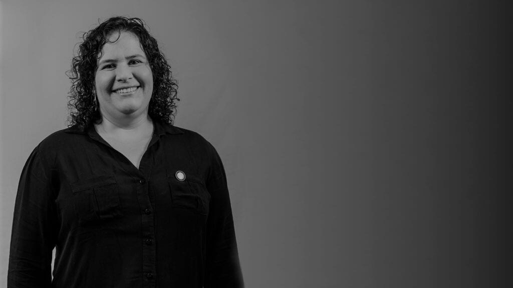 Mulher de pele clara, fios pretos cacheados na altura dos ombros sorrindo para foto. Ela veste blusa social preta de manha longa com botões no meio. É a dra. Paola Rueda.