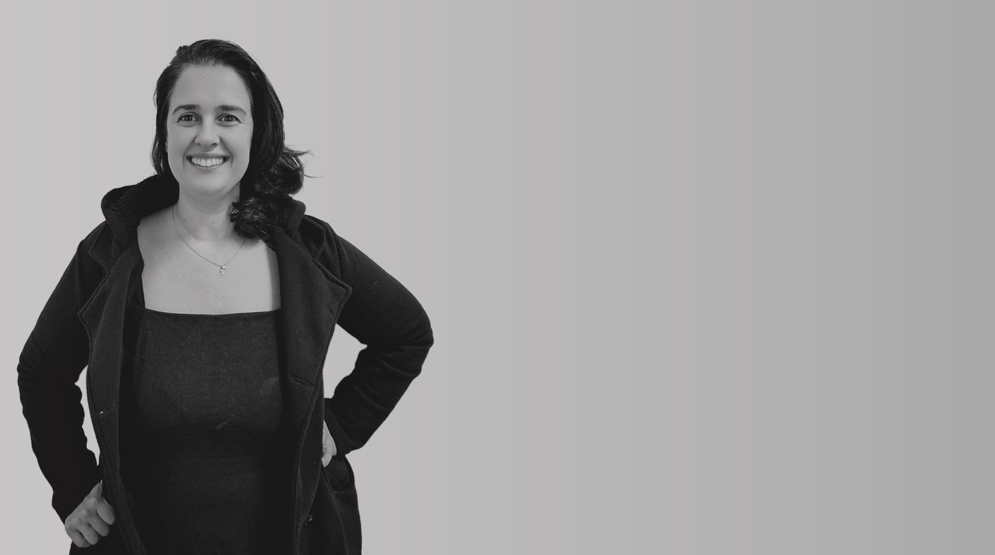 Mulher branca de fios pretos lisos e ondulados na ponta, em pé, sorrindo para foto. Em veste blusa e casaco pretos.