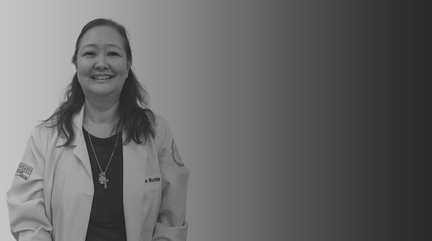 Mulher de traços orientais, cabelo preto liso com volume de tamanho médio sorri para foto. Ela veste jaleco branco de médica-veterinária, blusa preta e gargantilha prateada com um crucifixo na mesma cor. É a dra. Ayne Hayashi.