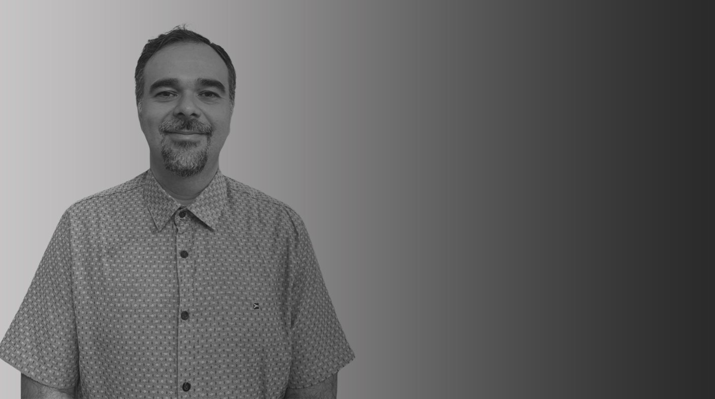 Homem branco de cavanhaque em pé sorrindo para foto. Ele veste blusa com estampa de pequenos riscos, mangas curtas e botões azuis marinho. Esse é Daniel Leite da Silva.