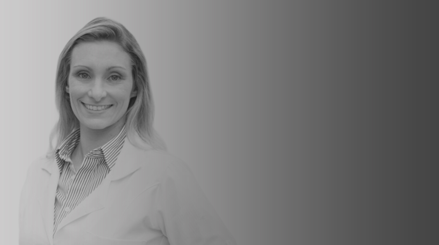 Mulher branca de fios loiros e lisos na altura dos ombros sorri para foto. Ela veste jaleco branco e blusa listrada azul e branca. É a dra. Maria Formenton.