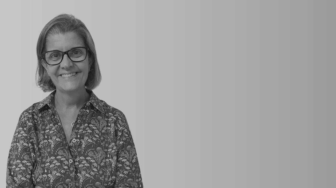 Mulher branca de fios lisos tamanho chanel sorri para foto. Ela usa óculos quadrado de armação preta e veste blusa estampada de mangas longas e botões. É Gisele Machado.