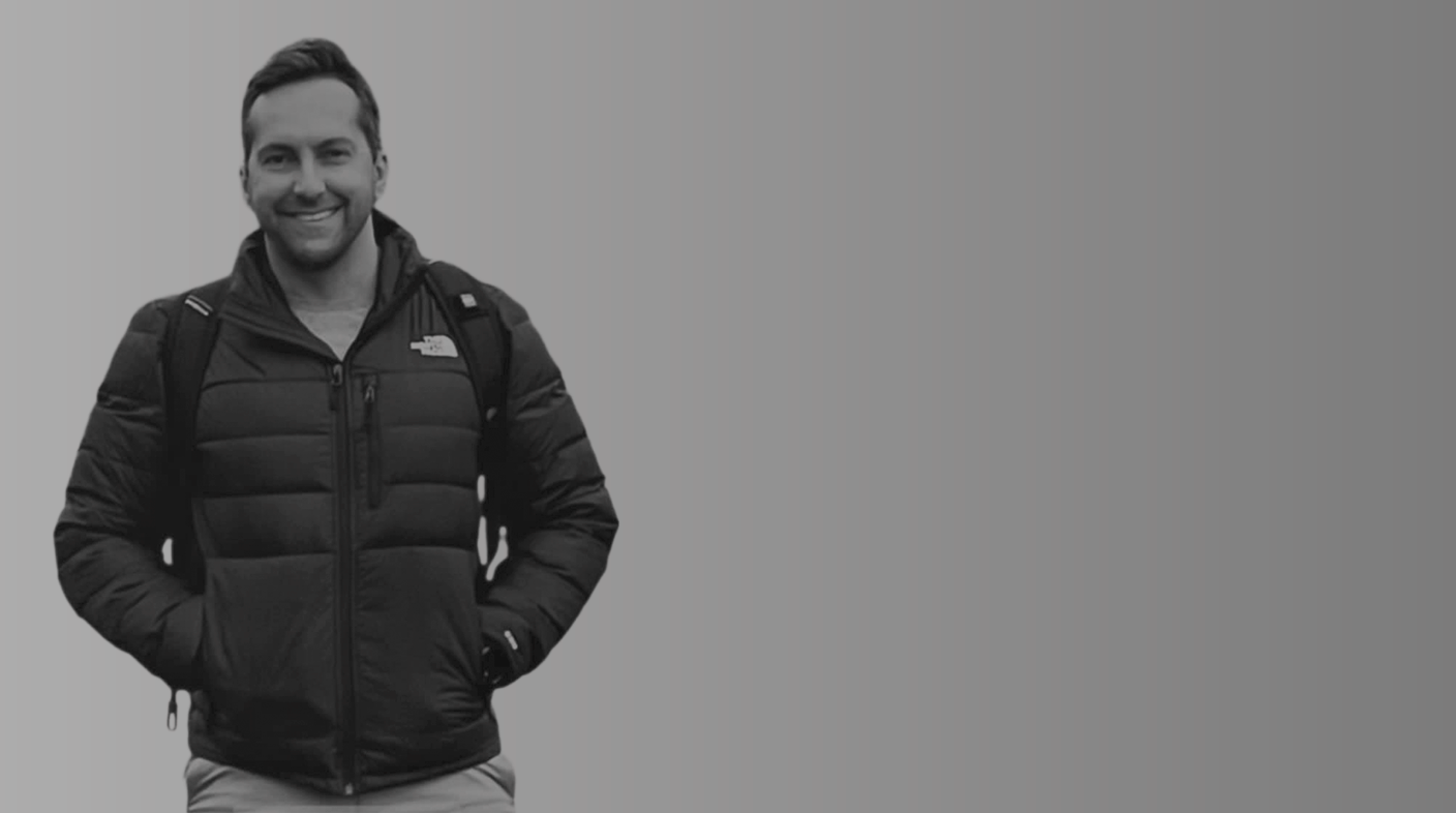 Homem branco jovem em pé sorrindo para foto. Ele usa casaco preto esportivo, está com uma mochila preta nas costas e mãos nos bolsos da blusa. É Marcos Roberto André.