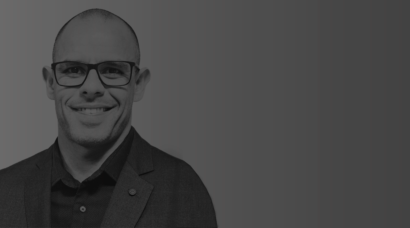 Homem branco careca sorri para foto. Ele usa óculos retangular grande de armação preta e veste blusa social de manga longa abotoada no meio. É o dr. Michel Luidge.