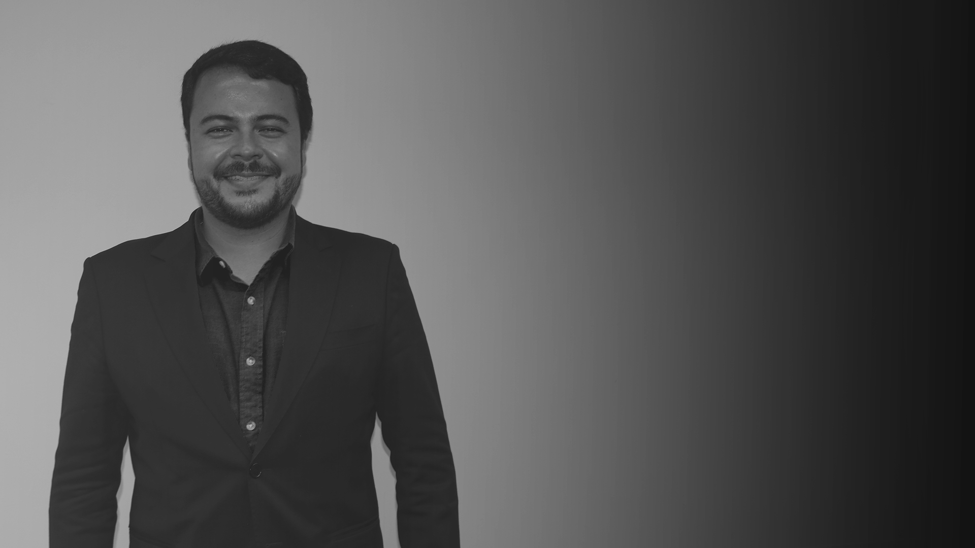 Homem branco em pé sorridente para foto. Ele veste terno escuro e blusa escura de botões brancos. Esse é Rodrigo Hayashi Goulart.