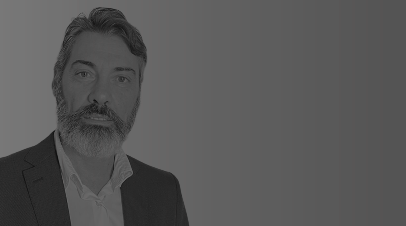 Homem branco de barba grisalha pousa sério para foto. Ele usa blazer escuro aberto e blusa branca social. É Rogério Amorim.