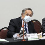 Presidente do CRMV-SP, Mário Eduardo Pulga, está sentando atrás da mesa de máscara branca e vestindo terno. Na mesa a bandeira do estado de São Paulo e uma placa com a identificação.