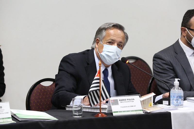 Presidente do CRMV-SP, Mário Eduardo Pulga, está sentando atrás da mesa de máscara branca e vestindo terno. Na mesa a bandeira do estado de São Paulo e uma placa com a identificação.