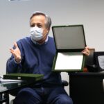 Presidente do CRMV-SP, Mário Eduardo Pulga, está sentado, de máscara e vestindo um suéter azul. Ele fala portando em mãos um estojo de veludo preto com uma placa metálica dentro