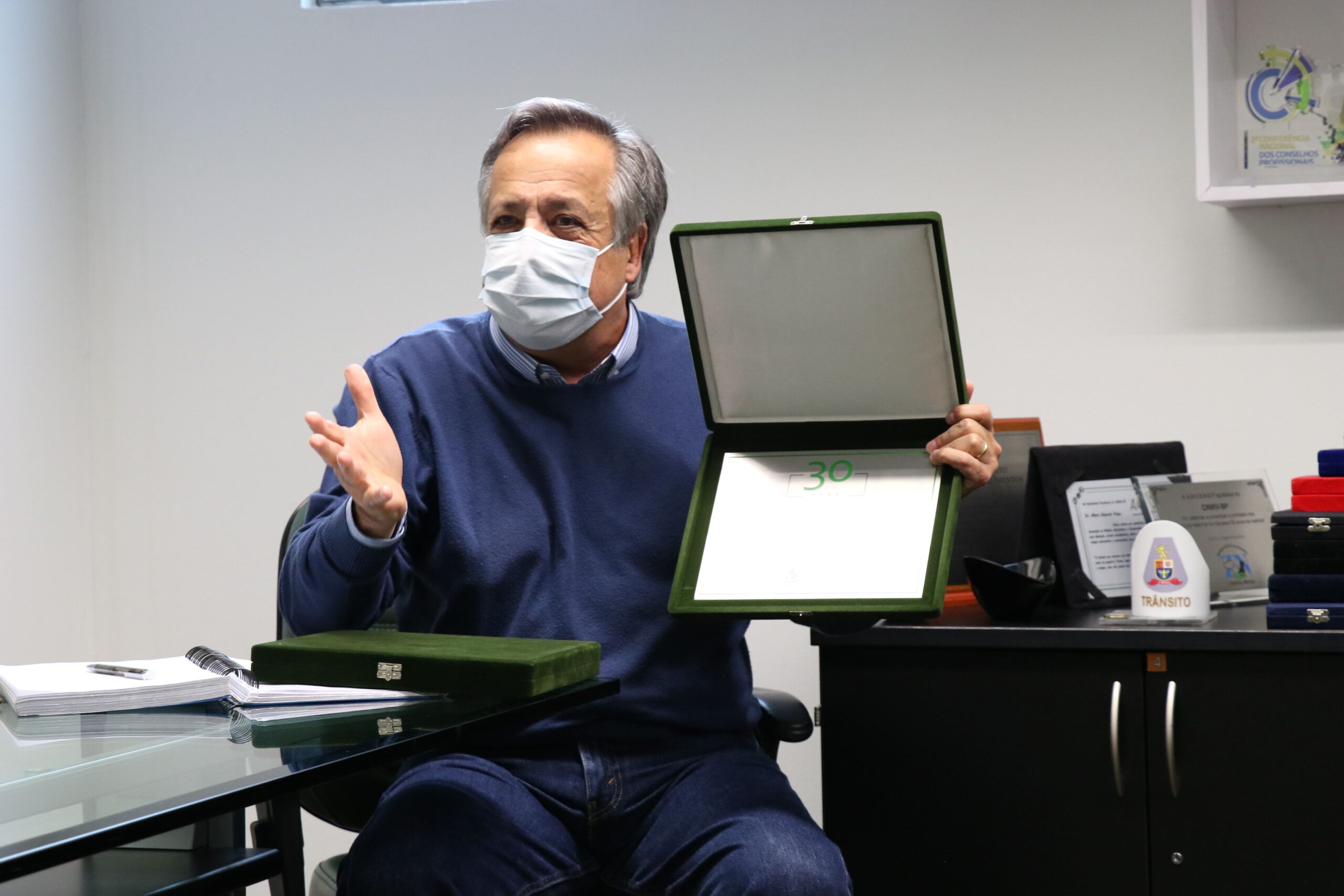 Presidente do CRMV-SP, Mário Eduardo Pulga, está sentado, de máscara e vestindo um suéter azul. Ele fala portando em mãos um estojo de veludo preto com uma placa metálica dentro