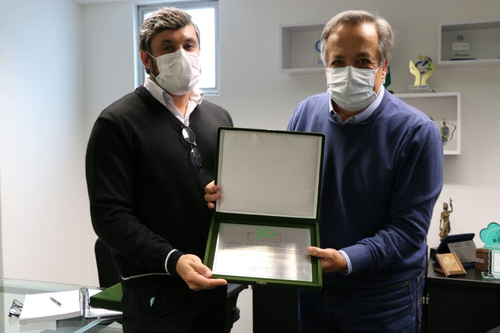 Presidente do CRMV-SP, Mário Eduardo Pulga entrega placa de homenagem ao funcionário Marcos Antonio. Ambos estão em pé, de máscara, e ambos posam segurando, um de cada lado, o estojo com a placa de homenagem em que é possível ler "30 anos"