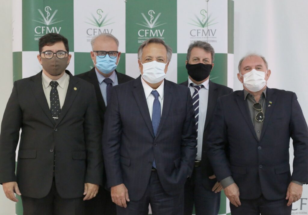 Membros da nova Câmara Técnica de Medicina Veterinária do CFMV possam, todos de terno e máscara, em frente ao painel quadriculado nas cores verde e branco e logo do Conselho Federal.