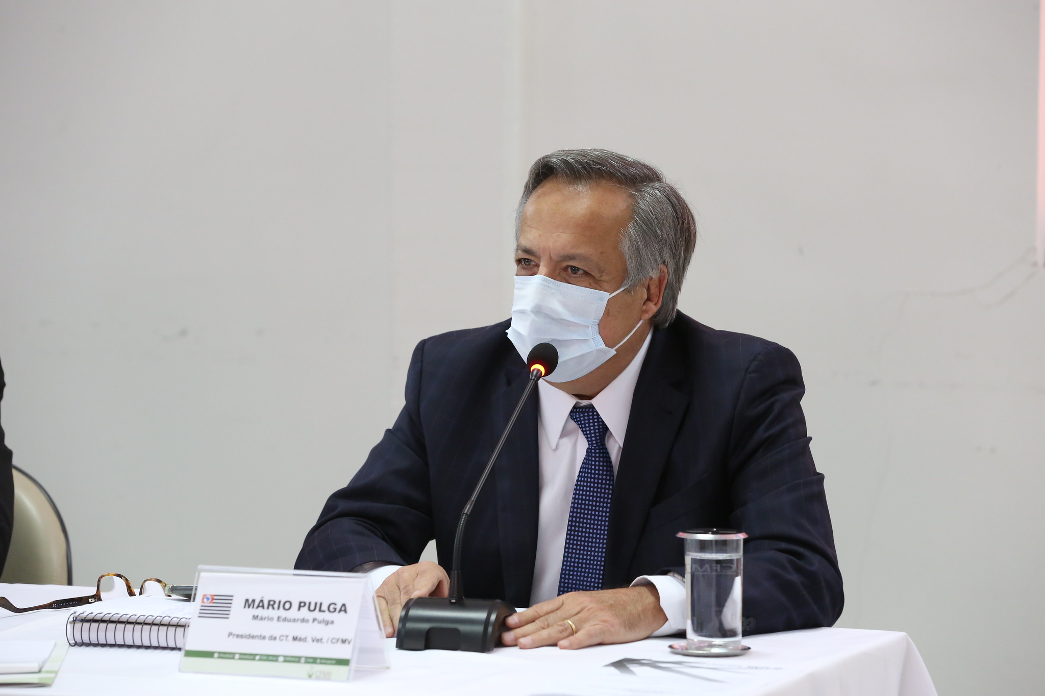 Mário Eduardo Pulga, de máscara, discursa sentado atrás de uma mesa em que estão placa com seu nome, microfone, e copo com água sob uma toalha branca.