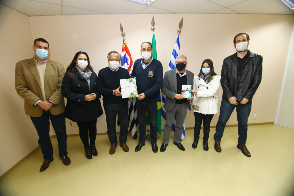 Em pé, em frente as bandeiras da cidade, do estado e do Brasil, estão da esquerda para a direita, o coordenador técnico médico-veterinário, Leonardo Burlini Soares; a secretária de Saúde, Margarete da Silva Correia; o presidente do Conselho, Mário Eduardo Pulga segurando o livro dos 50 anos da autarquia; o prefeito de São José dos Campos, Felicio Ramuth recebendo o livro; o vice-presidente do CRMV-SP, Odemilson Donizete Mossero segurando o Manual de RT; a diretora do departamento de Vigilância Epidemiológica, Mariana Keesen recebendo a publicação; e o gerente do Centro de Controle de Zoonoses (CCZ), Diego Amaral