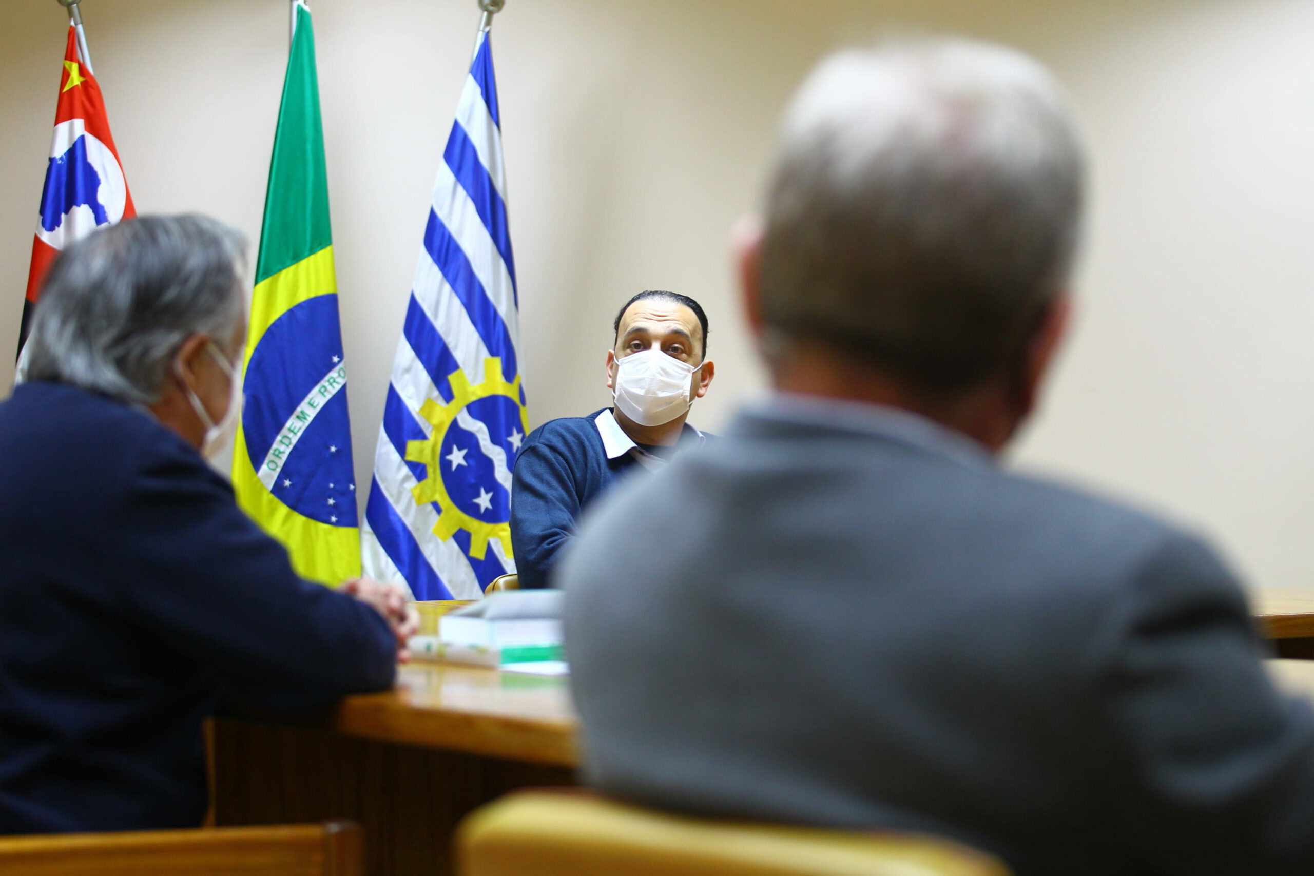 Prefeito de São José dos Campos sentado ao fundo em destaque em frente as bandeiras da cidade, do estado e do Brasil, e aparecem sentados de costas o presidente e o vice-presidente do CRMV-SP