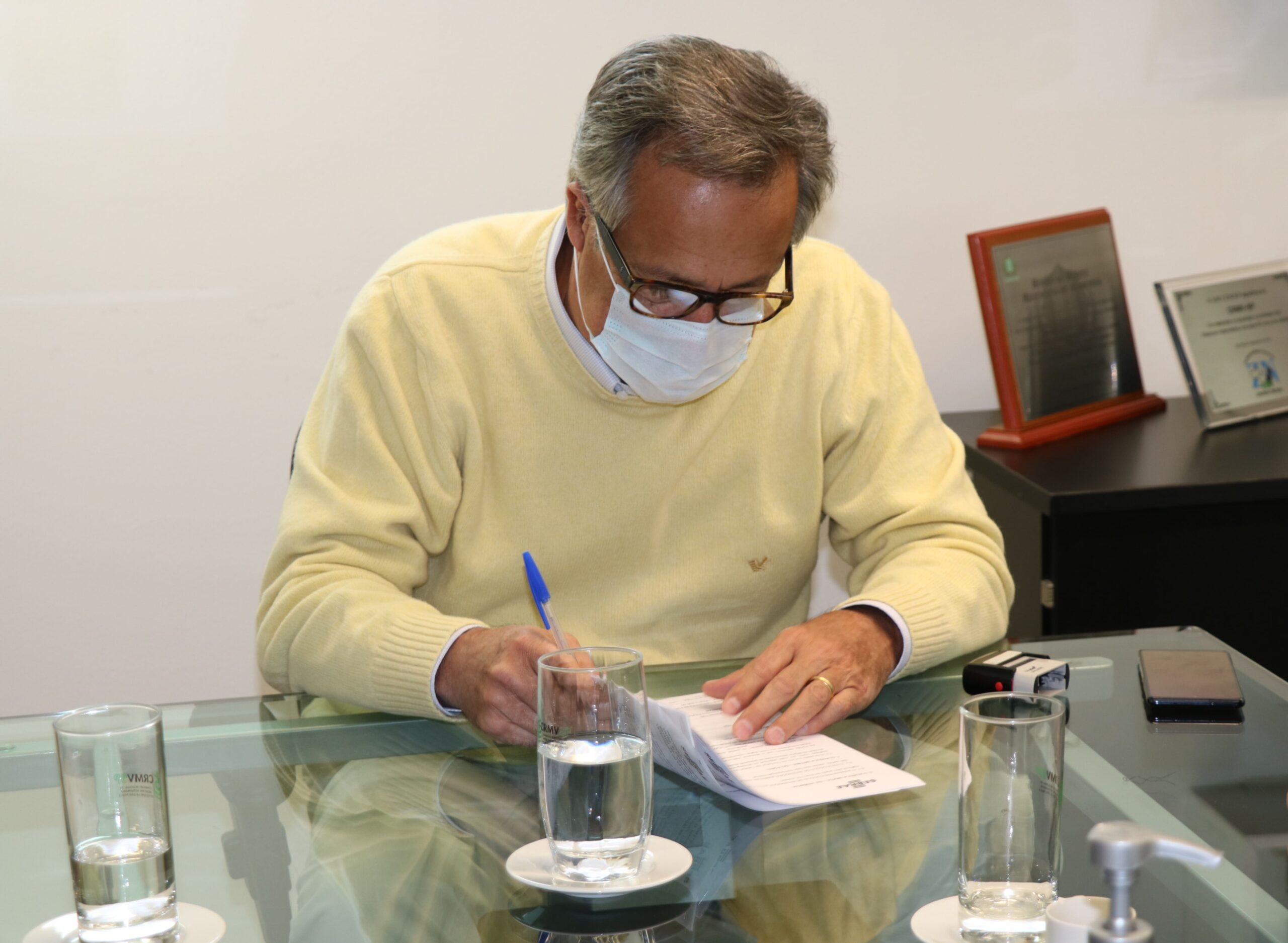 Presidente do CRMV-SP está de suéter bege, máscara e óculos, sentado à mesa assinando o protocolo de intenção.