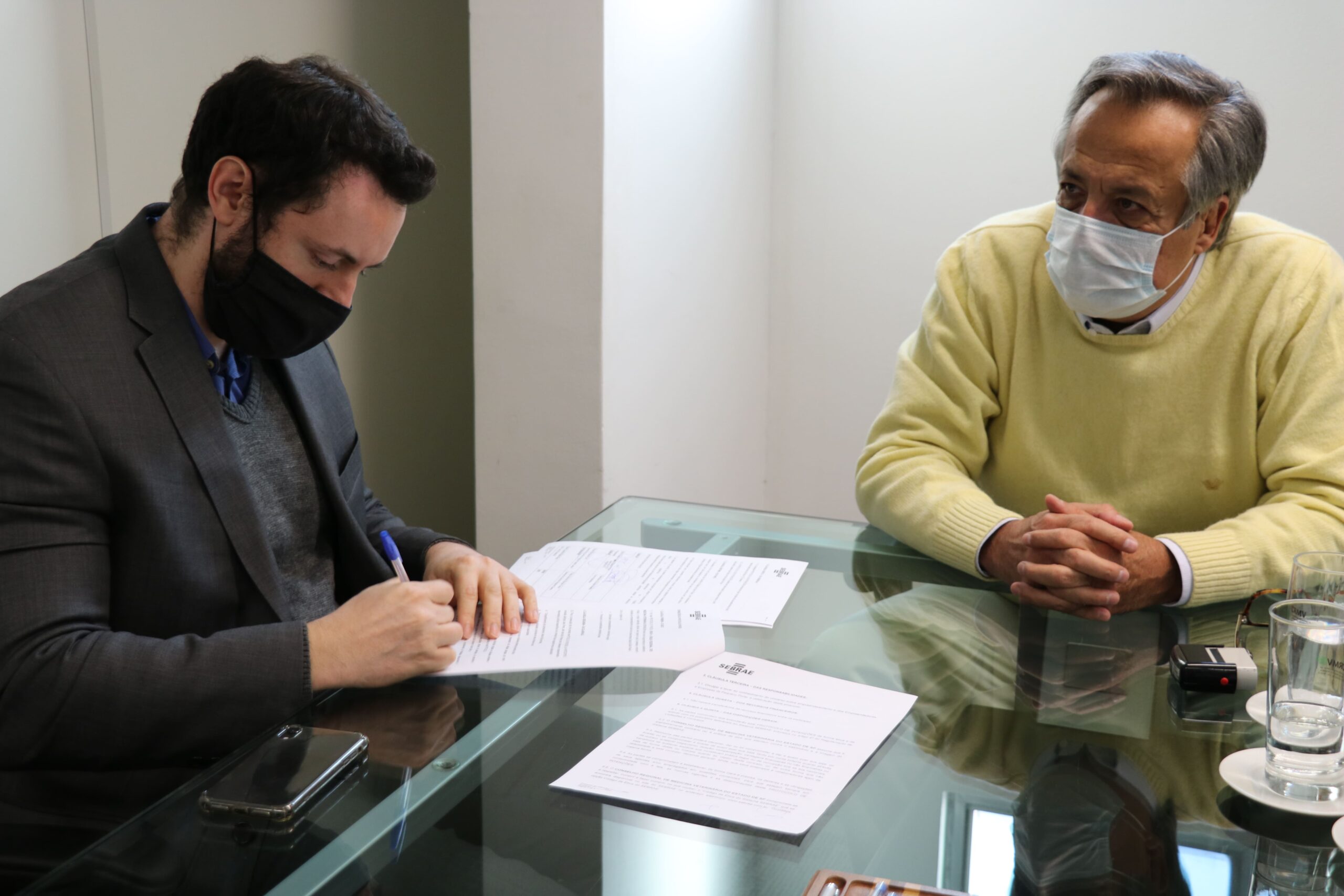 Presidente do CRMV-SP está de suéter bege, máscara e óculos, sentado à mesa olhando o gerente regional Capital leste do Sebrae, de máscara e casaco marrom escuro, assinar protocolo de intenção.