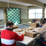 Membros da Comissão de Tomada de Contas estão sentados ao redor de uma mesa com diversas pastas e processos, e ao fundo janelas e o painel pantográfico xadrez verde e branco com a logomarca do CRMV-SP