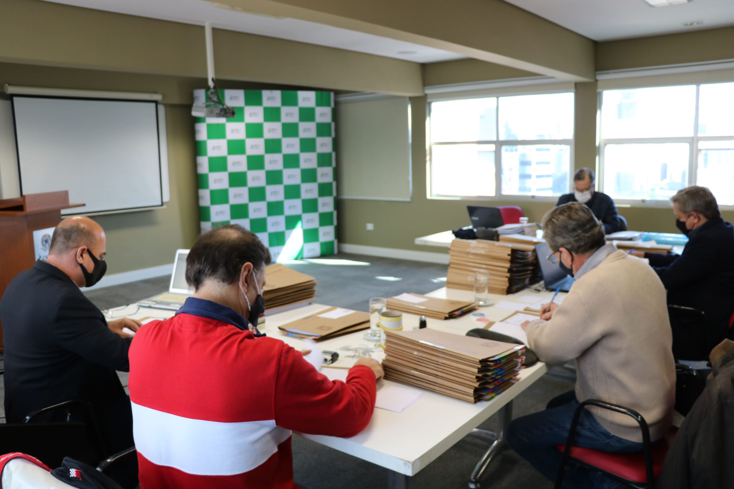 Membros da Comissão de Tomada de Contas estão sentados ao redor de uma mesa com diversas pastas e processos, e ao fundo janelas e o painel pantográfico xadrez verde e branco com a logomarca do CRMV-SP