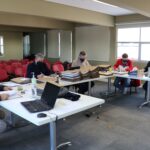 Membros da Comissão de Tomada de Contas sentados ao redes de mesas com pastas, processos e notebooks, ao fundo janelas e cadeiras vermelhas