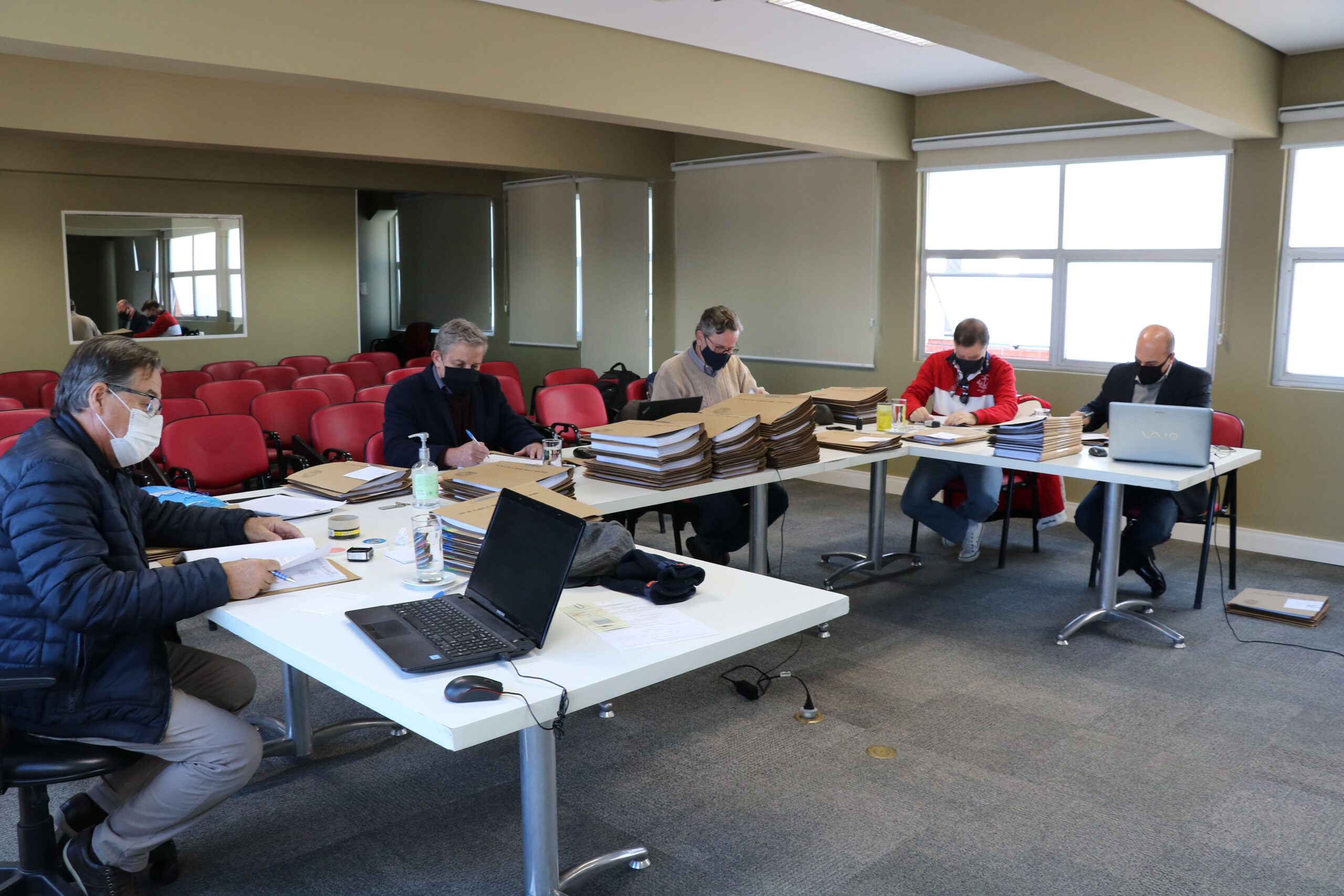 Membros da Comissão de Tomada de Contas sentados ao redes de mesas com pastas, processos e notebooks, ao fundo janelas e cadeiras vermelhas