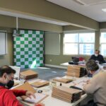 Membros da Comissão de Tomada de Contas estão sentados ao redor de uma mesa com diversas pastas e processos, e ao fundo janelas e o painel pantográfico xadrez verde e branco com a logomarca do CRMV-SP