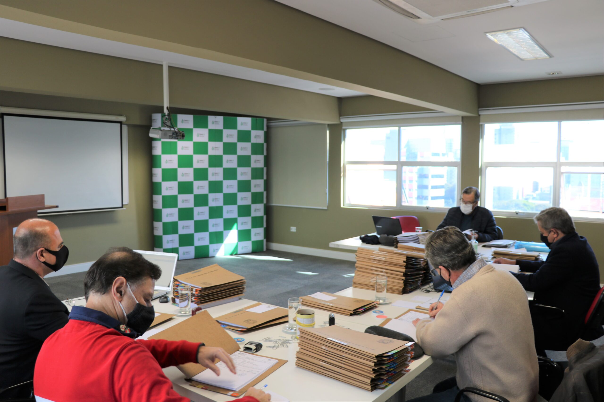 Membros da Comissão de Tomada de Contas estão sentados ao redor de uma mesa com diversas pastas e processos, e ao fundo janelas e o painel pantográfico xadrez verde e branco com a logomarca do CRMV-SP