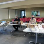 Membros da Comissão de Tomada de Contas sentados ao redes de mesas com pastas, processos e notebooks, ao fundo janelas e cadeiras vermelhas