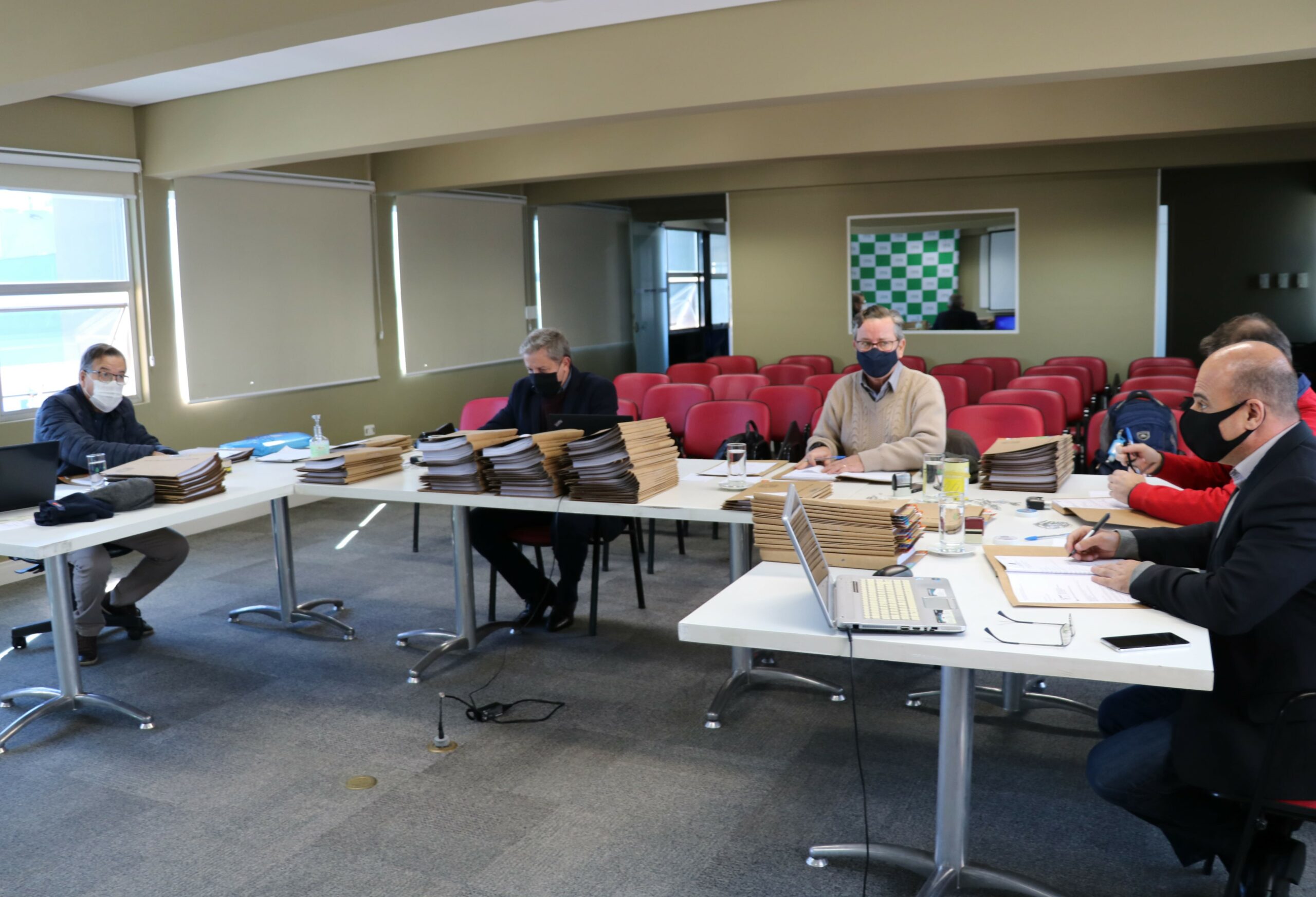 Membros da Comissão de Tomada de Contas sentados ao redes de mesas com pastas, processos e notebooks, ao fundo janelas e cadeiras vermelhas