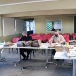 Membros da Comissão de Tomada de Contas sentados ao redes de mesas com pastas, processos e notebooks, ao fundo janelas e cadeiras vermelhas