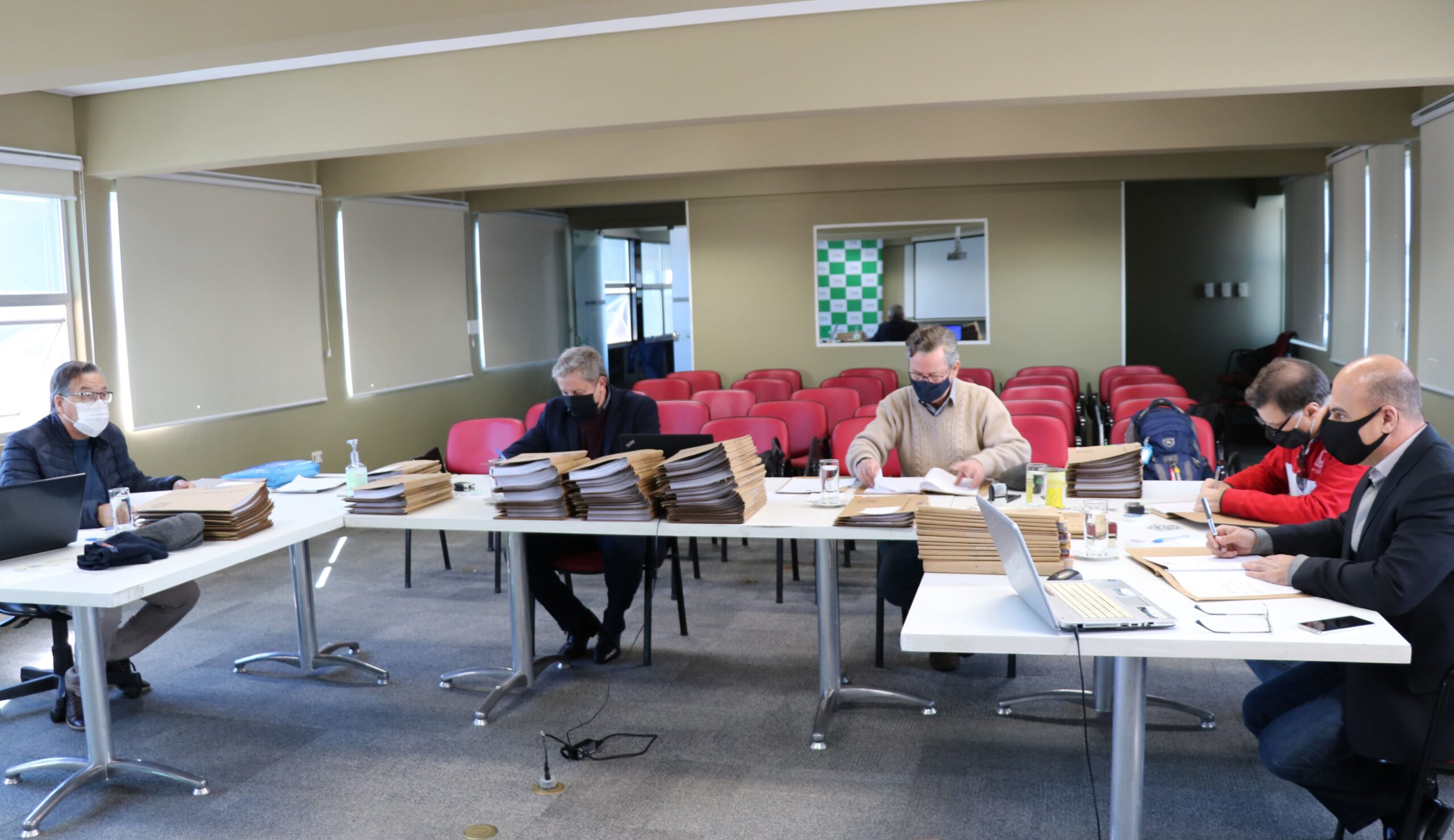 Membros da Comissão de Tomada de Contas sentados ao redes de mesas com pastas, processos e notebooks, ao fundo janelas e cadeiras vermelhas