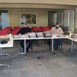 Membros da Comissão de Tomada de Contas sentados ao redes de mesas com pastas, processos e notebooks, ao fundo janelas e cadeiras vermelhas