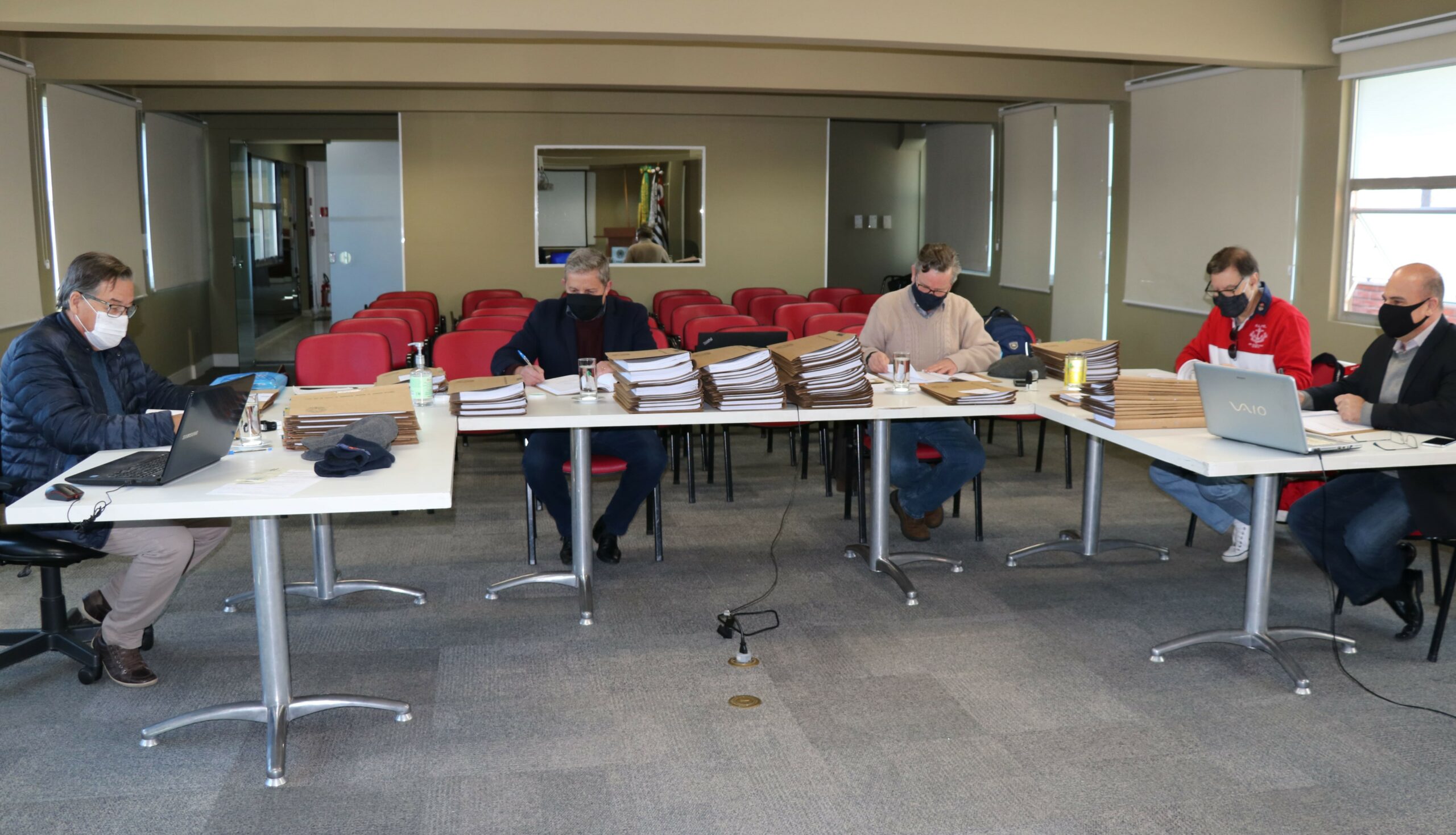 Membros da Comissão de Tomada de Contas sentados ao redes de mesas com pastas, processos e notebooks, ao fundo janelas e cadeiras vermelhas