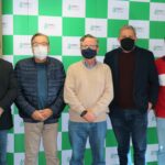 Integrantes da Comissão de Tomada de Contas de máscara possam de pé em frente ao painel pantográfico do CRMV-SP, mas cores verde e branco.