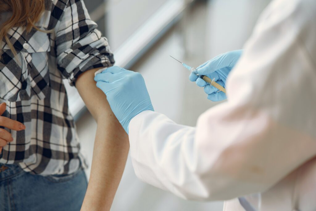 Mulher com camisa xadrez e jeans recebe vacina no braço de um profissional da saúde vestindo jaleco branco e luvas azuis