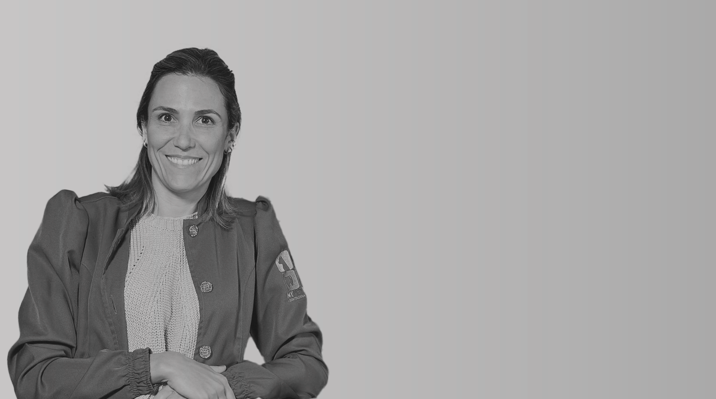 Mulher branca em pé sorrindo para foto. Ela usa jaleco de médica-veterinária, blusa clara e braços cruzados. É Mariana Lage Marques.