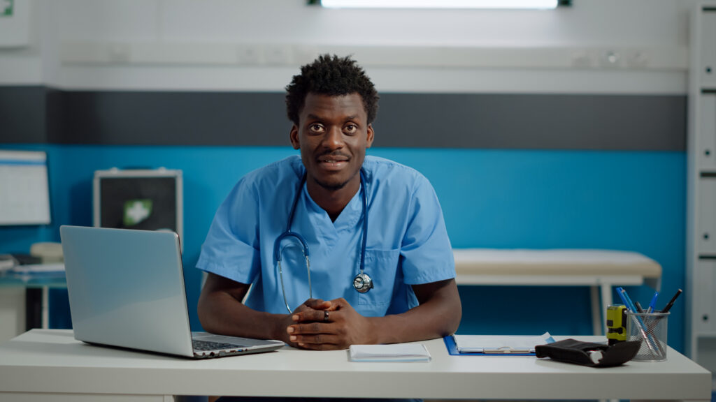 Médico-veterinário negro está sentado atrás de uma mesa com computador, olhando e sorrindo para a câmera.