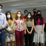 Grupo de oito pessoas em pé usando máscaras, entre eles uma mulher segura arranjo de flores.