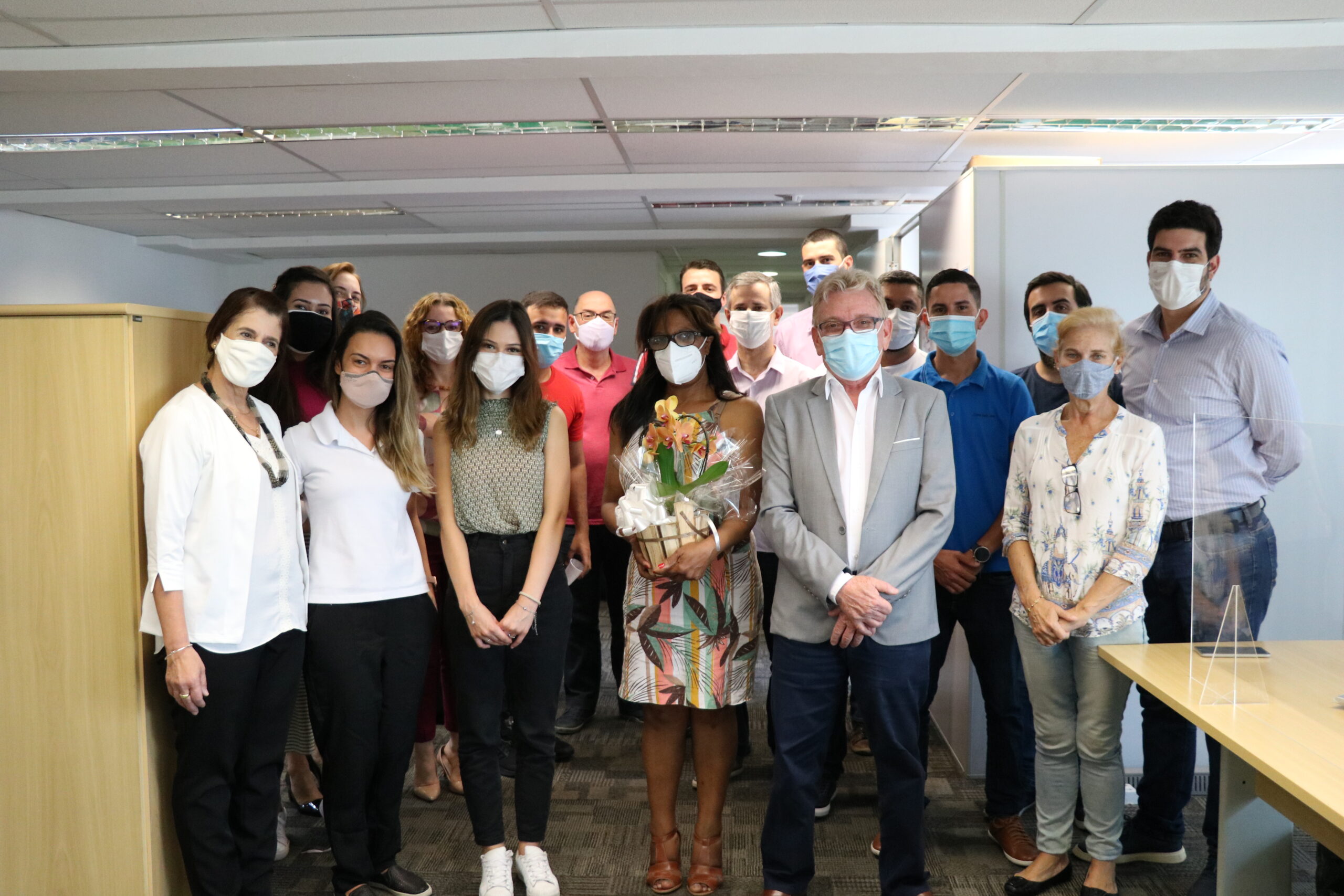 Dezoito pessoass em pé e com máscaras , Mulher no centro da imagem segura um arrajno de flores .