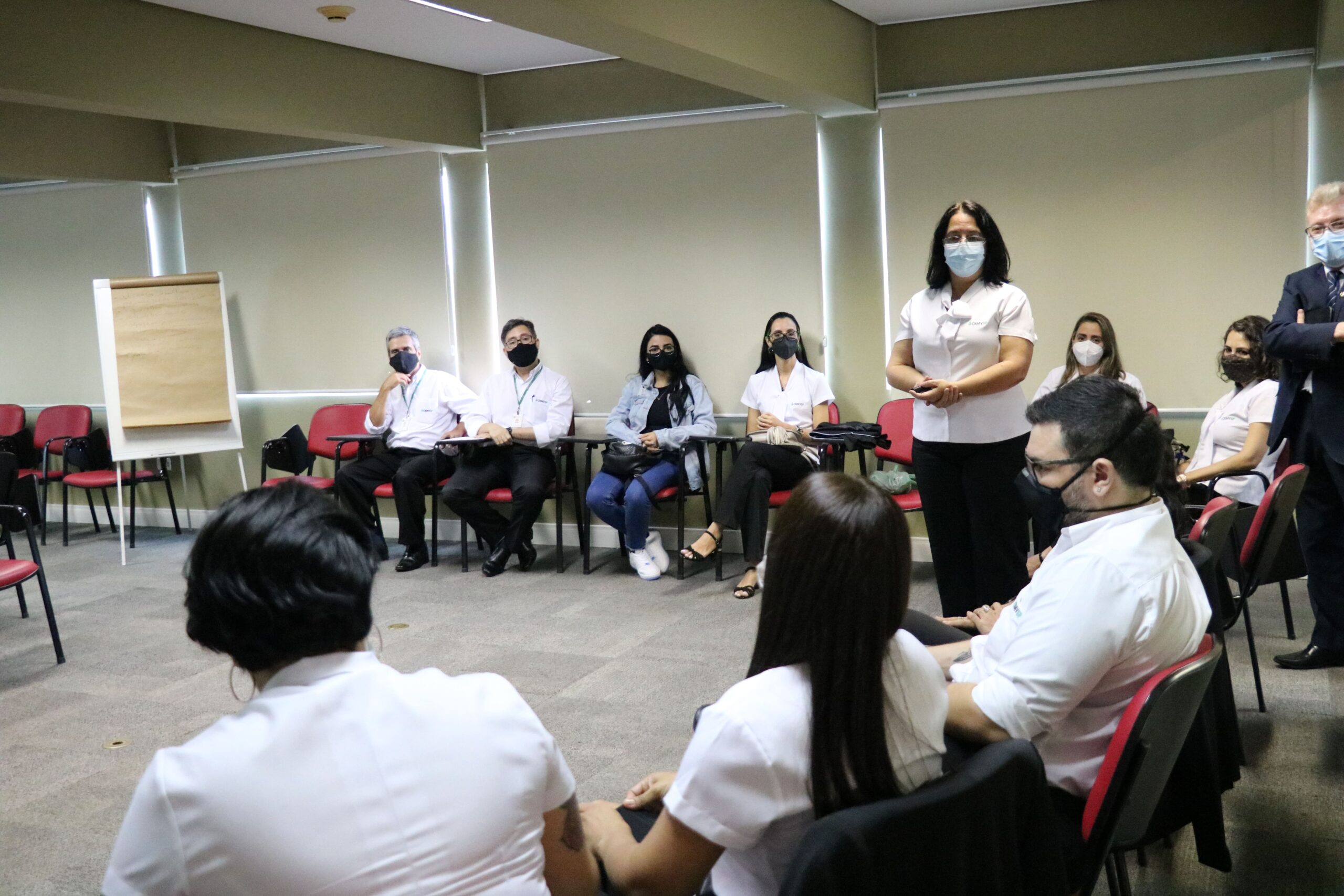 Funcionários das Unidades Regionais sentados de máscara sendo orientados por uma colaboradora do Conselho que está em pé.