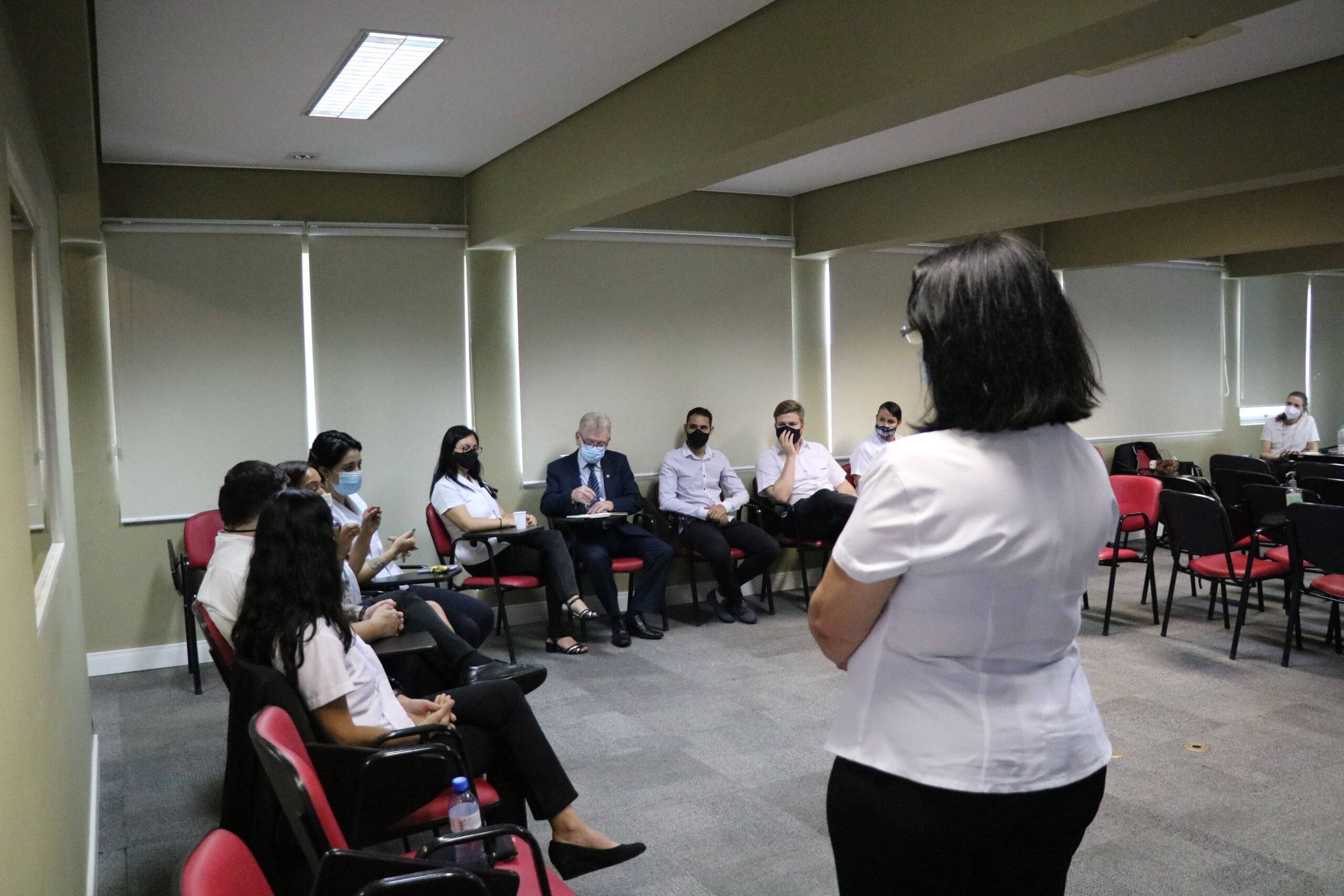 Colaboradora do Conselho está em pé, de costas para a câmera e de frente para os funcionários das Unidades Regionais do CRMV-SP sentados formando meia-lua e de máscaras. Entre eles está o presidnete do Conselho Odemilson Donizete Mossero.