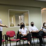 Á esquerda, em primeiro plano uma mulher em pé e de máscara faz orientação a três outros colaboradores sentados em semi-círculo à sua lateral.