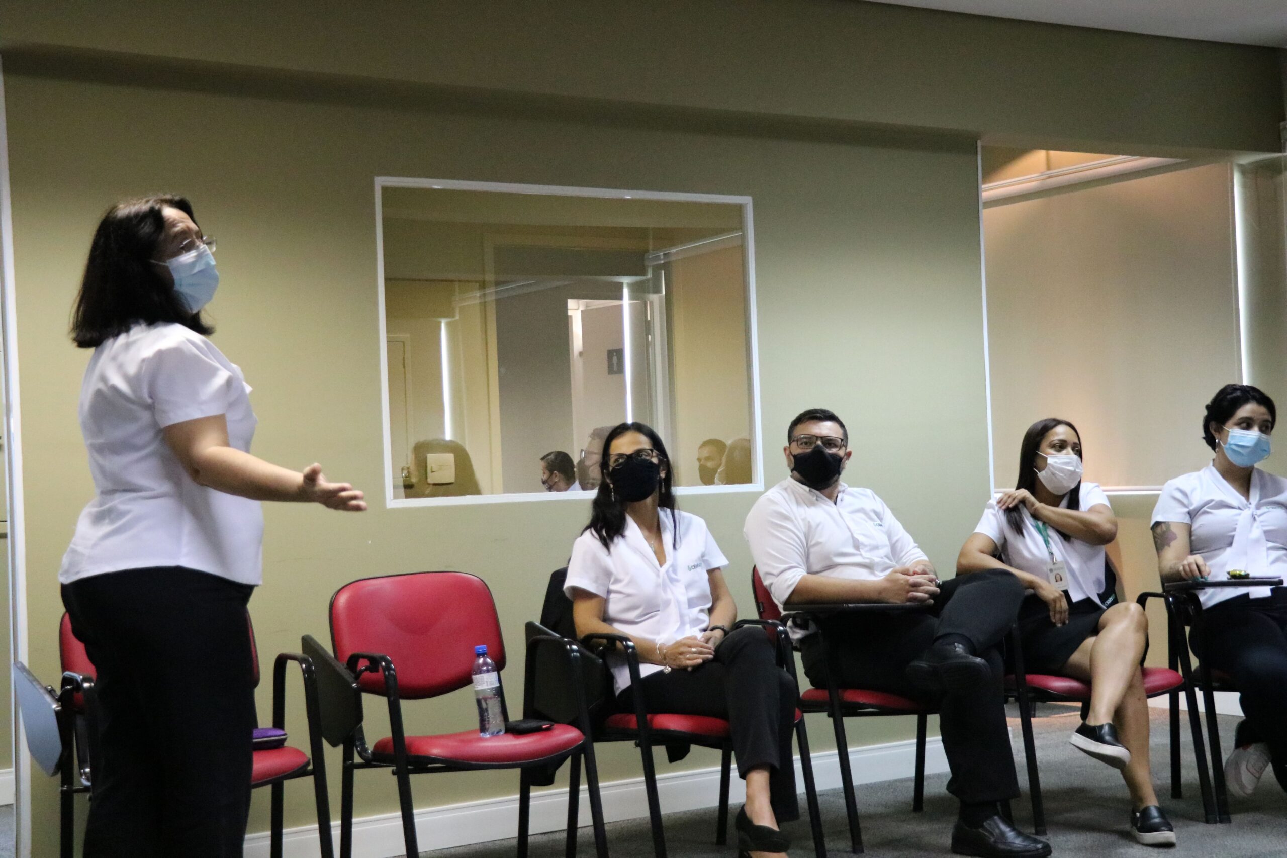 Á esquerda, em primeiro plano uma mulher em pé e de máscara faz orientação a três outros colaboradores sentados em semi-círculo à sua lateral.