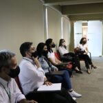 Seis pessoas sentada e usando máscaras estão encostados em uma parede branca.