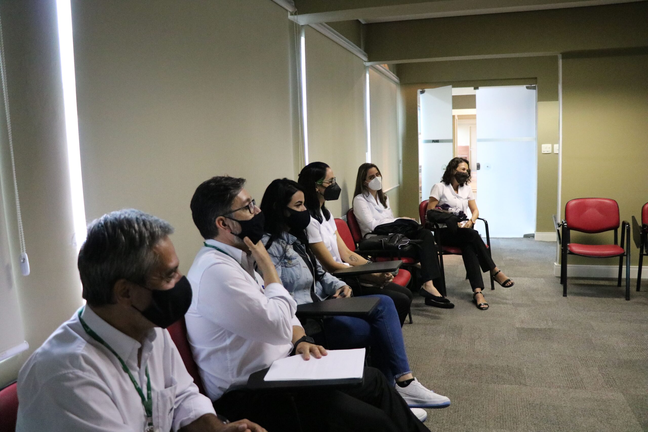 Seis pessoas sentada e usando máscaras estão encostados em uma parede branca.