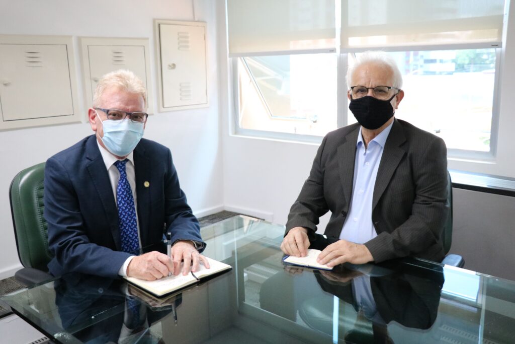 Na foto estão o presidente do CRMV-SP e o novo assessor de gabinete estão sentados diante de uma mesa de vidro posando para a foto. Ambos vestem paletó escuro e máscaras. Ao fundo, duas janelas abertas.