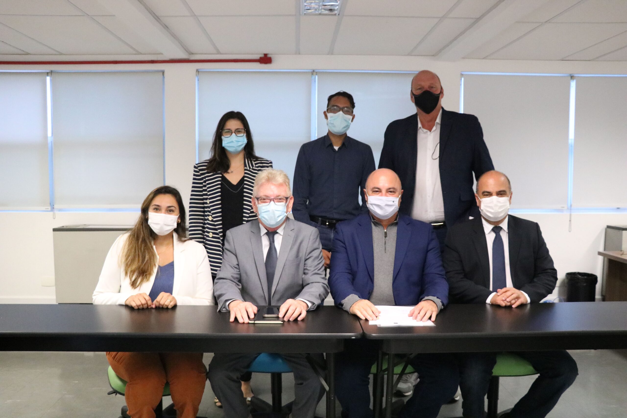 Na foto aparecem quatro pessoas sentadas e três atrás destas de pé. São duas mulheres e cinco homens. Todos estão de máscara e posam para a foto.