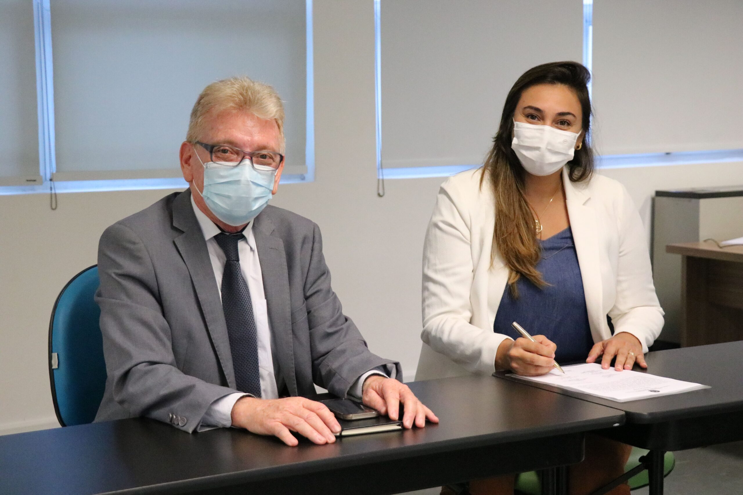 Na foto estão sentados lado a lado e de máscara, posando para a câmera, o presidente do CRMV-SP e a zootecnista Thalita Oliveira Cucki, que faz a menção de assinar o termo de posse como integrante da Comissão de Nutrição Animal.