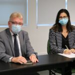 Na foto estão sentados lado a lado e de máscara, posando para a câmera, o presidente do CRMV-SP e a médica-veterinária Brana Sanctos, que faz a menção de assinar o termo de posse como integrante da Comissão de Nutrição Animal.